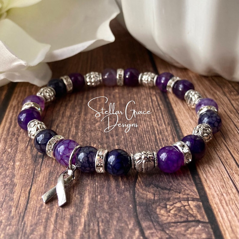 May include: Purple and silver beaded bracelet with a silver ribbon charm. The bracelet features a variety of purple agate beads and silver spacer beads. The ribbon charm is a symbol of support and awareness.