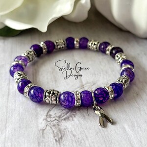 May include: Purple agate gemstone bracelet with silver accents and a purple ribbon charm. The bracelet is made with small, round beads and has a delicate, feminine look.