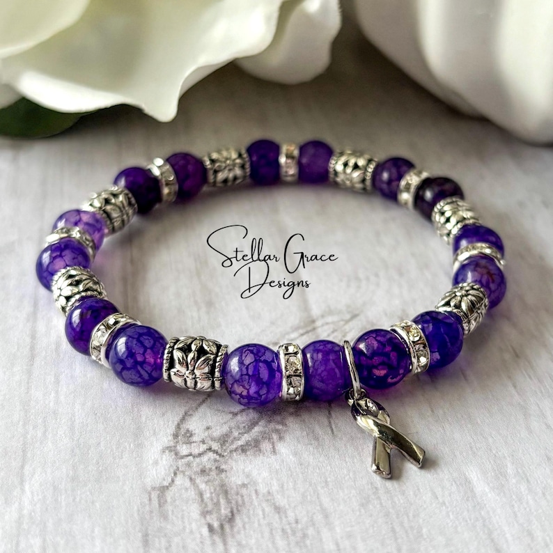 May include: Purple agate gemstone bracelet with silver accents and a purple ribbon charm. The bracelet is made with small, round beads and has a delicate, feminine look.