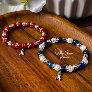May include: Two beaded bracelets featuring silver-toned spacers and charms. One bracelet consists of red beads, while the other features alternating blue and white crackle beads. The text Stellar Grace Designs is visible on the blue and white bracelet.