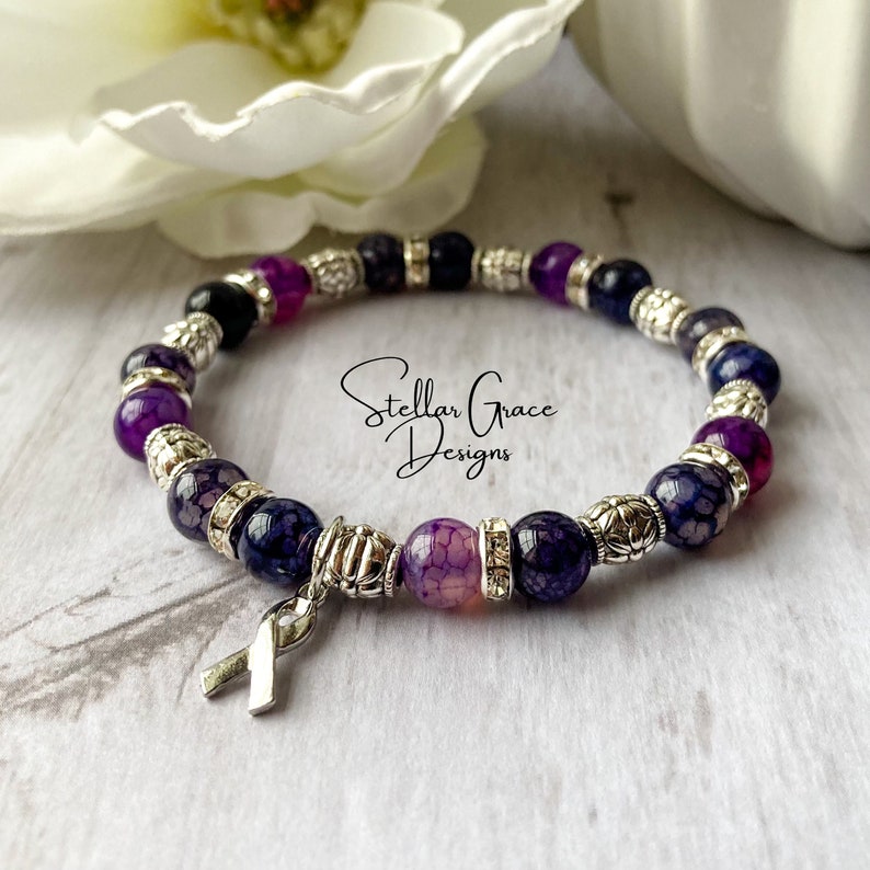 May include: Purple and silver beaded bracelet with a silver ribbon charm. The bracelet features a variety of purple and silver beads, including agate and metal beads. The ribbon charm is a symbol of support and awareness.