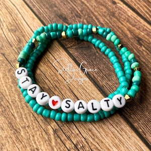 May include: Teal and green beaded bracelet with the words "Stay Salty" and a red heart charm.