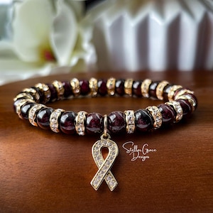May include: A beaded bracelet with dark red beads and gold-colored accents. A gold-colored ribbon charm with clear crystals hangs from the bracelet. The bracelet is on a brown surface.