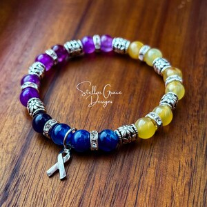 May include: A multi-colored beaded bracelet with a silver ribbon charm. The bracelet features purple, blue, and yellow beads with silver accents. The bracelet is on a wooden surface. Stellar Grace Designs.