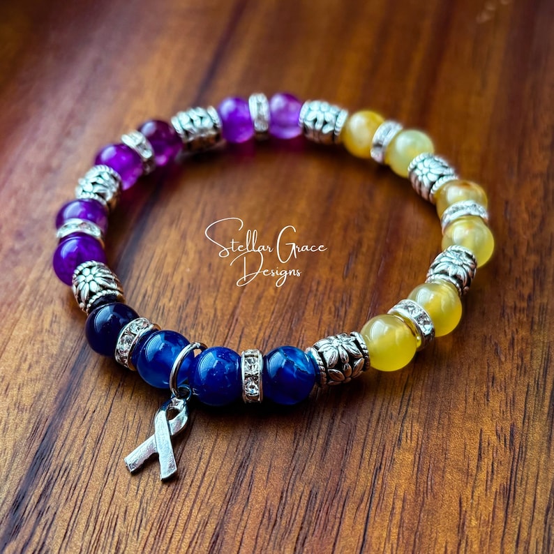 May include: A multi-colored beaded bracelet with a silver ribbon charm. The bracelet features purple, blue, and yellow beads with silver accents. The bracelet is on a wooden surface. Stellar Grace Designs.