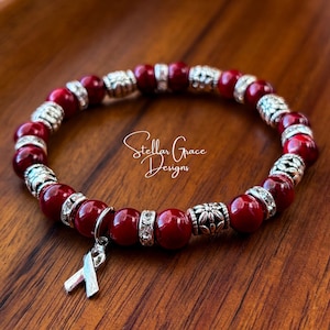 May include: Red beaded bracelet with silver accents and a silver ribbon charm. The bracelet is on a wooden surface. Stellar Grace Designs.