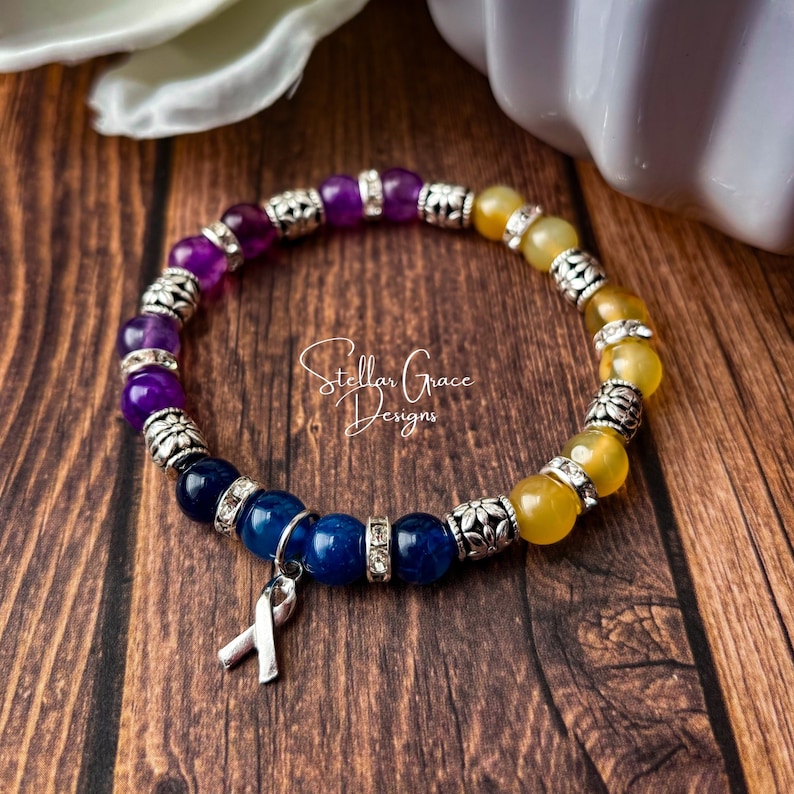 May include: A purple, blue, and yellow beaded bracelet with silver accents and a silver ribbon charm. The bracelet is on a wooden surface. Stellar Grace Designs.