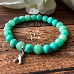 May include: A teal green beaded bracelet with a silver ribbon charm. The bracelet is on a wooden surface. Stellar Grace Designs is written on the surface.