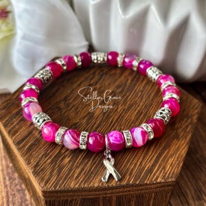 May include: A stretch bracelet featuring round, vibrant pink agate beads with white banding, interspersed with silver-toned metal spacers and clear rhinestone accents. A silver-toned awareness ribbon charm hangs from the center of the bracelet.
