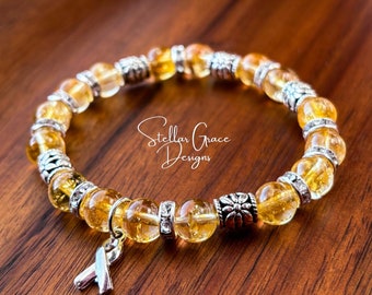 May include: A beaded bracelet featuring translucent yellow citrine-colored round beads alternating with silver-toned metal spacers and floral accents. A silver-toned awareness ribbon charm hangs from the bracelet, which displays the text Stellar Grace Designs.