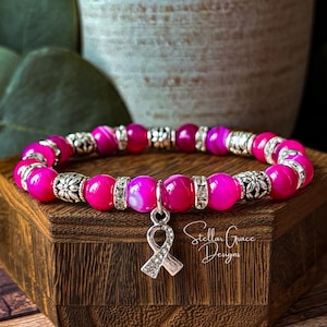 May include: A stretch bracelet featuring vibrant hot pink beads, silver-toned floral spacers, and clear rhinestone accents. A silver-toned awareness ribbon charm encrusted with small clear rhinestones hangs from the center of the bracelet.