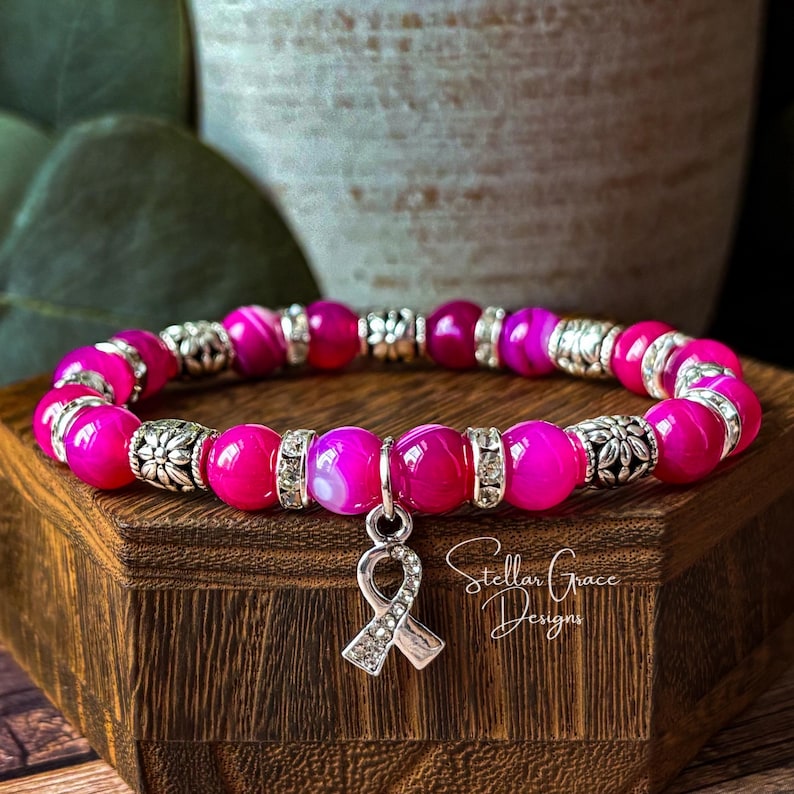 May include: A stretch bracelet featuring vibrant hot pink beads, silver-toned floral spacers, and clear rhinestone accents. A silver-toned awareness ribbon charm encrusted with small clear rhinestones hangs from the center of the bracelet.