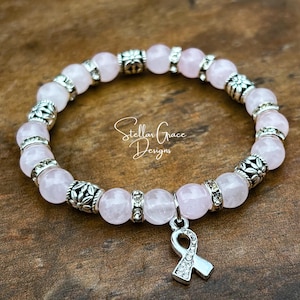 May include: A stretch bracelet featuring pale pink round beads, silver-toned metal spacers with floral patterns, and small clear crystal accents. A silver-toned awareness ribbon charm with clear crystals hangs from the bracelet. Text reads Stellar Grace Designs.