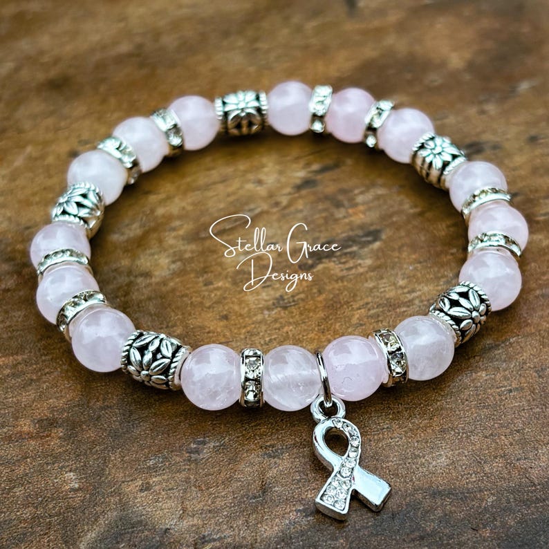 May include: A stretch bracelet featuring pale pink round beads, silver-toned metal spacers with floral patterns, and small clear crystal accents. A silver-toned awareness ribbon charm with clear crystals hangs from the bracelet. Text reads Stellar Grace Designs.