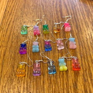 May include: A collection of 14 pairs of gummy bear dangle earrings. The earrings are made of clear resin and feature a variety of colours, including pink, blue, green, yellow, orange, and purple.