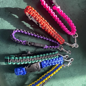 May include: A collection of colorful paracord wrist straps in pink, red, purple, green, blue, and yellow. Each strap features a black plastic buckle and a silver metal clip for attaching keys or other items.