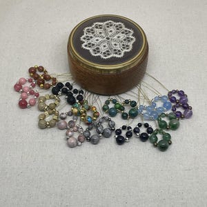 May include: A collection of beaded rings in various colors, including pink, brown, black, gray, blue, green, and purple, are displayed around a wooden box with a lace design on the lid. The rings are strung on thin, clear cords.