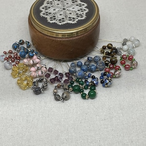 Puede incluir: Una caja de baratijas de madera con un borde de latón y un diseño de encaje en la tapa. Anillos de cuentas variados en varios colores, incluyendo azul, verde, rosa y amarillo, están dispuestos alrededor de la caja.