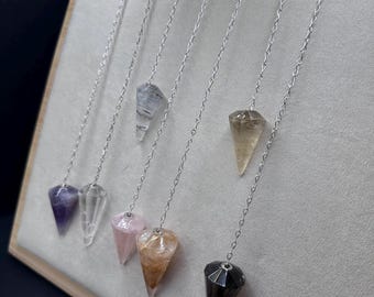 Puede incluir: Una colección de péndulos de cristal en varios colores, incluyendo púrpura, transparente, rosa y marrón, cada uno suspendido por una cadena plateada. Los péndulos se exhiben sobre una superficie clara e inclinada, con un fondo oscuro.