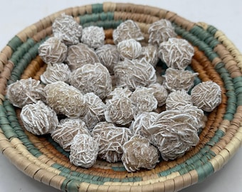 May include: A collection of desert rose crystals, with a textured, layered appearance, displayed in a woven basket with green, tan, and brown stripes. The crystals have a light brown colour with white accents.