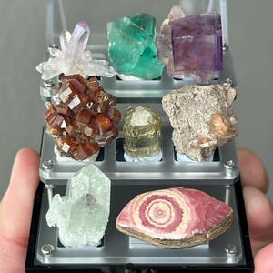 May include: A clear acrylic display case with nine compartments, each containing a different type of mineral specimen. The minerals include amethyst, fluorite, green calcite, red garnet, green apatite, a grey rock, a pink and white banded stone, and a clear quartz crystal.