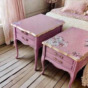 May include: Two matching pink bedside tables with gold accents and floral designs. The tables have two drawers each and are made of wood.