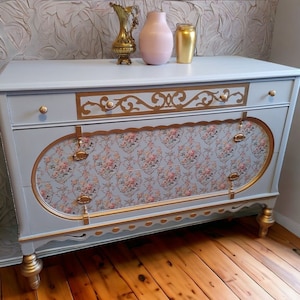 Peut inclure: Une commode bleu clair avec des accents dorés et des tiroirs à motifs floraux. La commode a quatre pieds et repose sur un sol en bois.