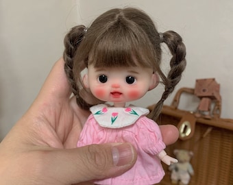 May include: A small doll with brown pigtails, wearing a pink dress with a white collar featuring a floral design. The doll has rosy cheeks and is wearing pink shoes. The doll is held in a hand.