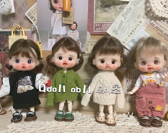 May include: Four dolls with different outfits and hairstyles are displayed. The dolls have fair skin, rosy cheeks, and large eyes. They wear a black dress, a green sweater, a white sweater, and pink overalls. The text "Qdoll obl 오비츠" is visible.