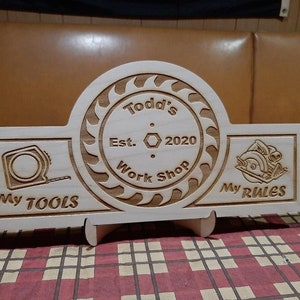 May include: A wooden sign with the text "Todd's Work Shop Est. 2020" in a circle with a saw blade design. The sign also has the text "My Tools" and "My Rules" on either side of the circle.