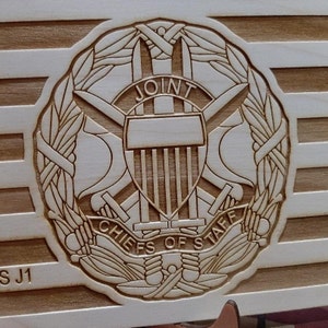 Puede incluir: Una placa de madera con una imagen grabada con láser del sello de los Jefes del Estado Mayor Conjunto. El sello presenta una corona de laurel, espadas cruzadas y las palabras "Joint Chiefs of Staff".