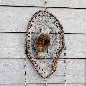 May include: A brown and white dreamcatcher with beads hanging from the bottom. The dreamcatcher is made of natural materials and has a small piece of fur in the center. The dreamcatcher is hanging on a light gray wall with the text "THEREBELTREE.COM" on the wall.