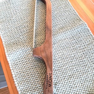 May include: A wooden cheese slicer with a curved handle and a stainless steel wire. The handle is engraved with "SNOW WOODWORK". The slicer is resting on a light green textured surface.