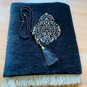 May include: A black prayer rug with an ornate gold and white design, a black tassel, and a cream-colored fringe. A black beaded rosary rests on the rug. The rug is folded on a wooden surface.
