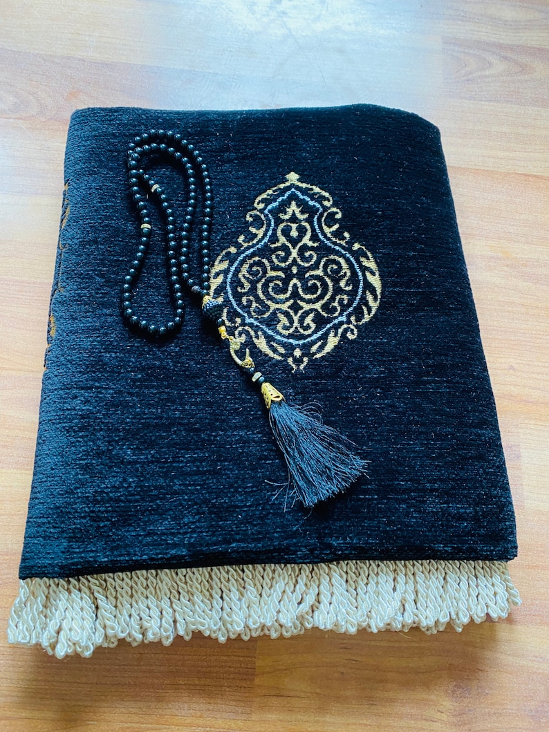 May include: A black prayer rug with an ornate gold and white design, a black tassel, and a cream-colored fringe. A black beaded rosary rests on the rug. The rug is folded on a wooden surface.