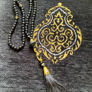 May include: Black prayer beads with a tassel and a decorative gold accent, resting on a black prayer rug with a gold and white ornate design. The beads are strung together, and the tassel is long and flowing.
