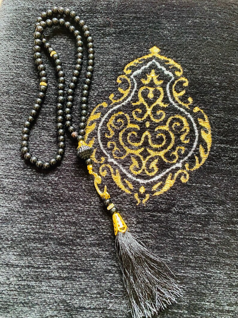 May include: Black prayer beads with a tassel and a decorative gold accent, resting on a black prayer rug with a gold and white ornate design. The beads are strung together, and the tassel is long and flowing.