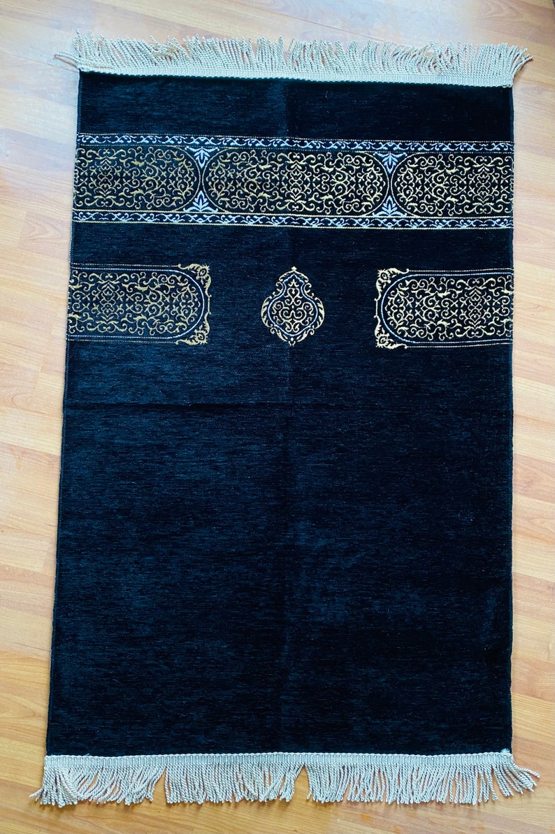 May include: A black prayer rug with gold-colored ornate designs. The rug has a rectangular shape with a decorative border and fringe at the ends. The design includes a central medallion and two rectangular panels.
