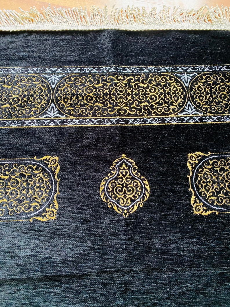 May include: Black prayer rug with gold and white ornate designs. The rug features a central medallion and decorative borders with intricate patterns. The edges are finished with a cream-colored fringe.