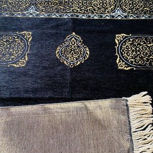 May include: Black prayer rug with gold and white ornate designs. The rug features a detailed border and decorative medallions. The bottom edge has a fringe. The rug is made of a soft, textured material.