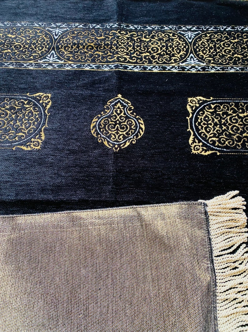 May include: Black prayer rug with gold and white ornate designs. The rug features a detailed border and decorative medallions. The bottom edge has a fringe. The rug is made of a soft, textured material.