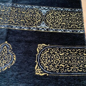 May include: Black prayer rug with gold and white ornate designs. The rug features a detailed border and decorative patterns. The top edge has a cream-colored fringe.