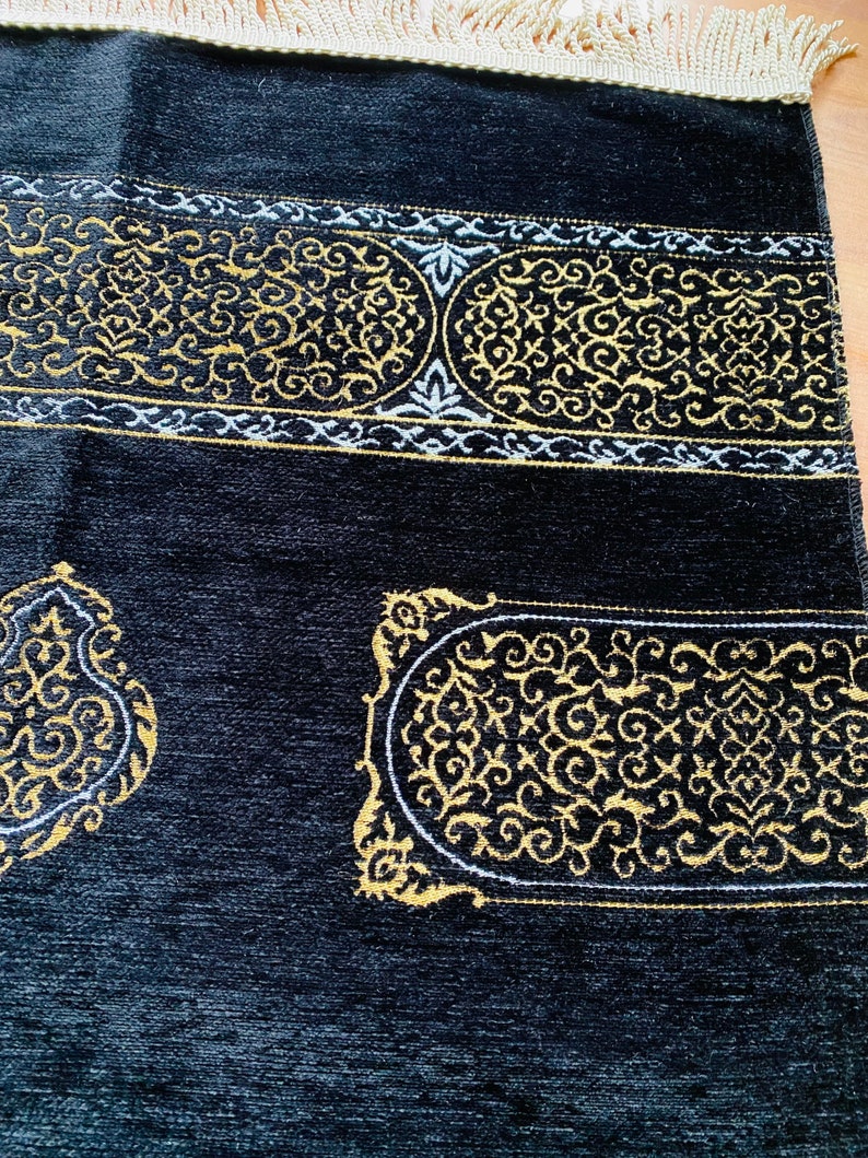 May include: Black prayer rug with gold and white ornate designs. The rug features a detailed border and decorative patterns. The top edge has a cream-colored fringe.