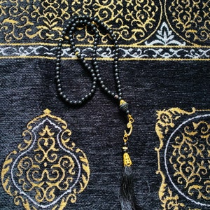 May include: A black prayer bead necklace with a tassel, resting on a black prayer rug with gold and white ornate patterns. The beads are small and round. The rug has a textured surface.