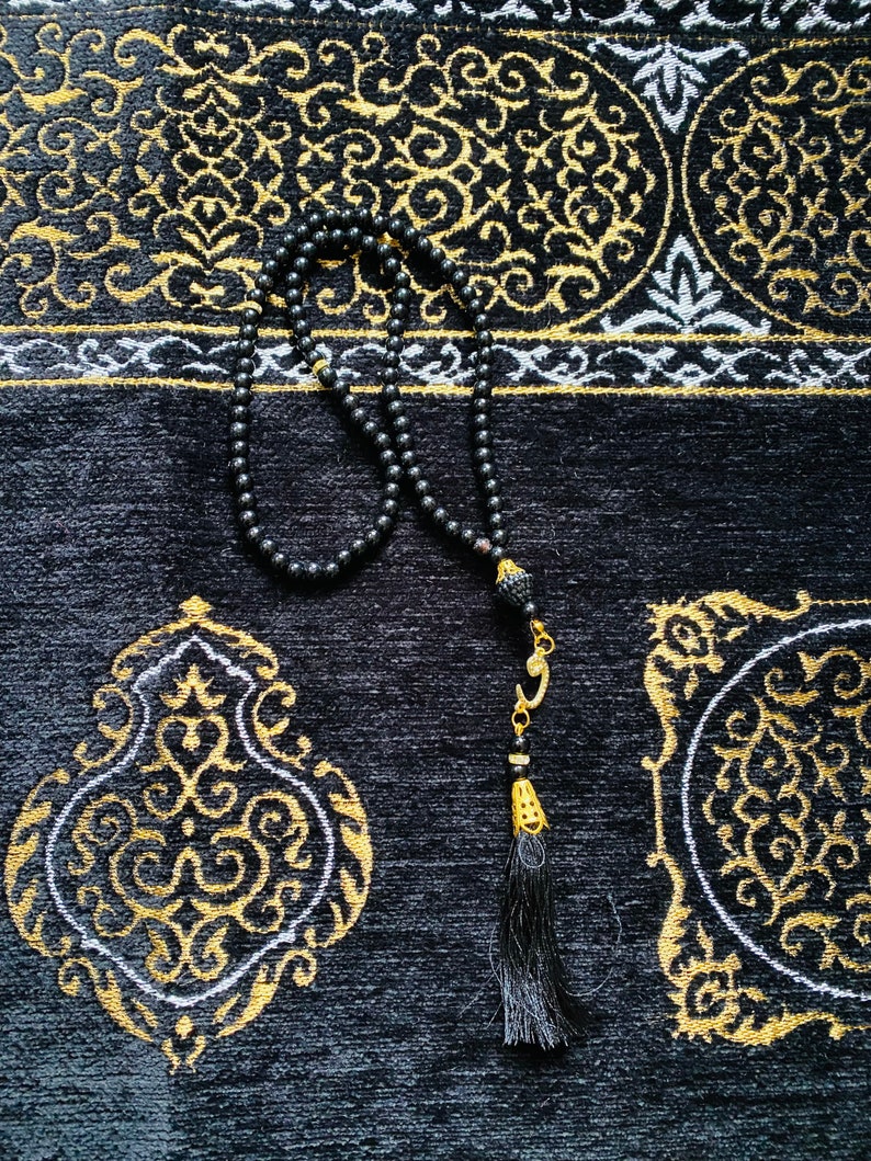 May include: A black prayer bead necklace with a tassel, resting on a black prayer rug with gold and white ornate patterns. The beads are small and round. The rug has a textured surface.