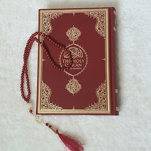 以下が含まれることがあります： 金色の装飾が施された、タイトルが「THE HOLY QUR'AN」のバーガンディ色の本と、お揃いのビーズロザリオ。ロザリオにはタッセルと装飾的なチャームが付いています。本とロザリオは、白くテクスチャ加工された表面に表示されています。