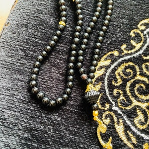 May include: A black prayer bead necklace with a gold-colored decorative element and a tassel. The beads are small and round, and the necklace is resting on a black prayer rug with gold-colored patterns.