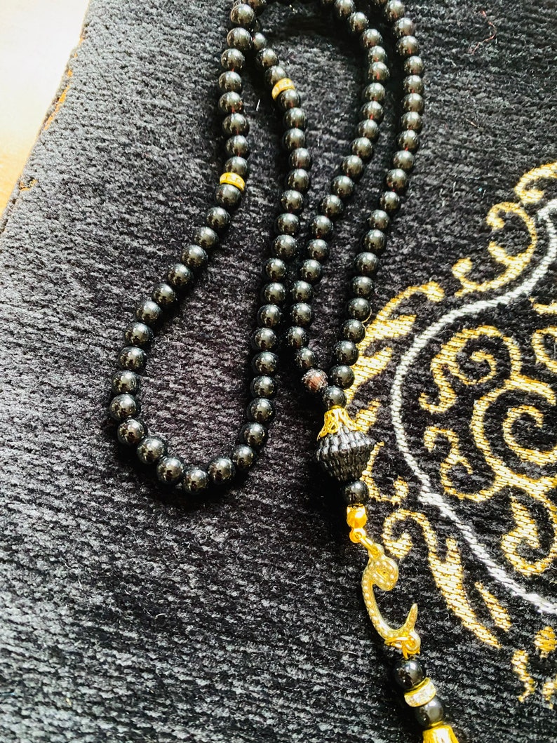 May include: A black prayer bead necklace with a gold-colored decorative element and a tassel. The beads are small and round, and the necklace is resting on a black prayer rug with gold-colored patterns.