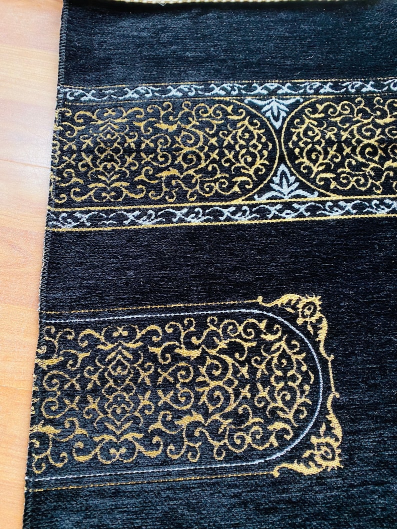 May include: Black prayer rug with gold and white ornate designs. The rug features a central rectangular design with a curved top and bottom, and a border with intricate patterns. The rug is made of a soft, plush material.