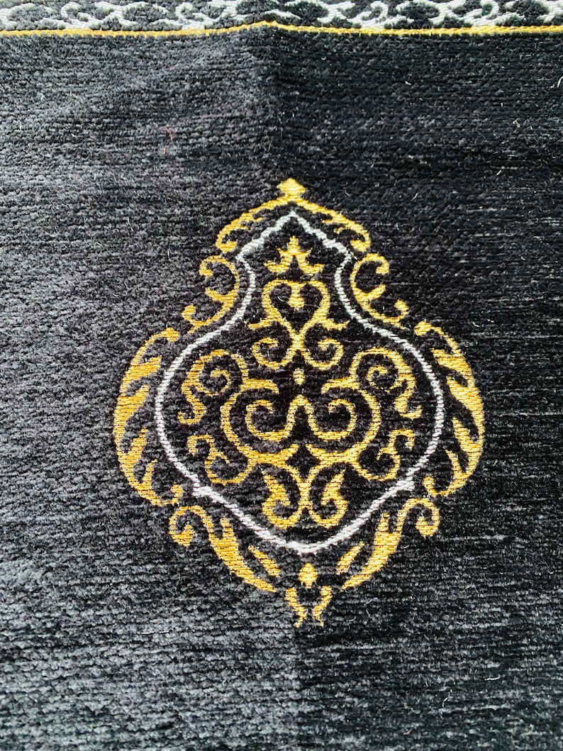May include: Black prayer rug with an ornate gold and white design. The central motif is a stylized, symmetrical shape with swirling patterns. The rug has a textured surface and a decorative border.